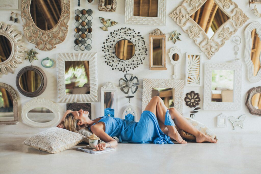 An adult woman in a blue dress relaxes indoors among artistic wall mirrors.